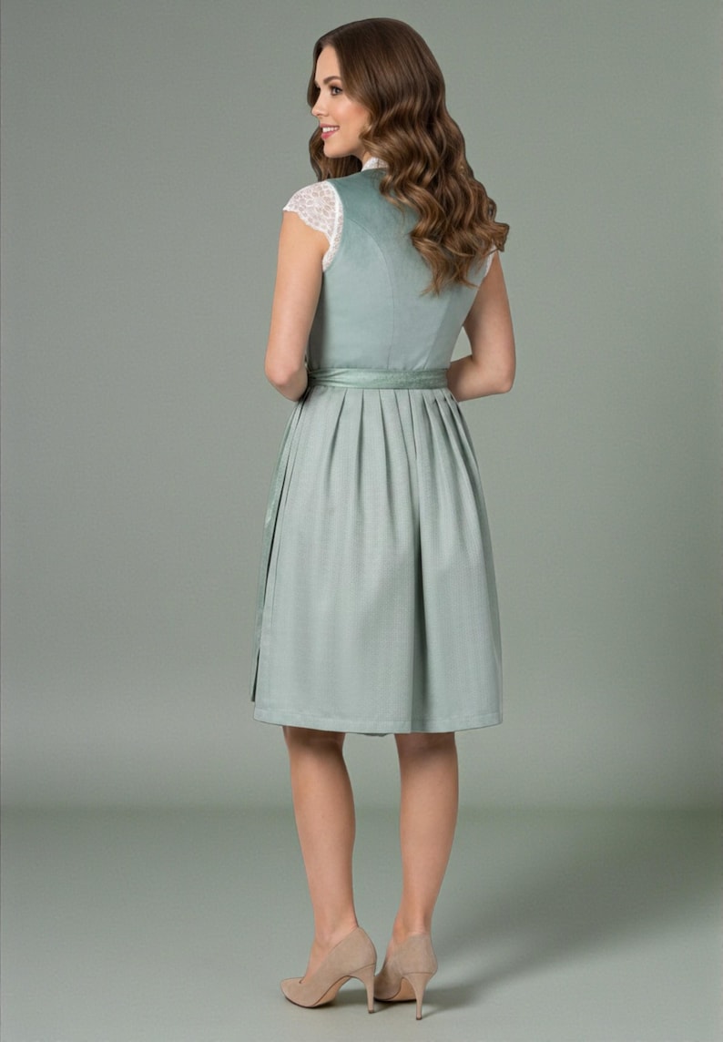 K&ouml;nnte beinhalten: Ein salbeigr&uuml;nes Dirndl mit &auml;rmellosem Oberteil, hochgeschlossenem Ausschnitt und einem Faltenrock. Das Kleid wird mit einer wei&szlig;en Spitzenbluse mit kurzen &Auml;rmeln, einem passenden Satin-Taillenband und beigen Pumps kombiniert.