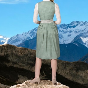 May include: A woman wearing a traditional green dirndl dress with white lace sleeves and a white blouse. The dress has a patterned design and a matching belt. The background features a mountain range and a clear blue sky.