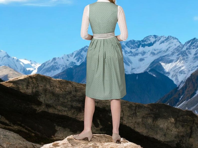 May include: A woman wearing a traditional green dirndl dress with white lace sleeves and a white blouse. The dress has a patterned design and a matching belt. The background features a mountain range and a clear blue sky.