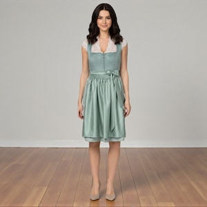 K&ouml;nnte beinhalten: Ein knielanges Dirndl in Salbeigr&uuml;n mit strukturiertem Stoff, Frontrei&szlig;verschluss und einer wei&szlig;en Spitzenbluse darunter. Das Kleid enth&auml;lt eine passende salbeigr&uuml;ne Sch&uuml;rze, die mit einer Schleife in der Taille gebunden ist.