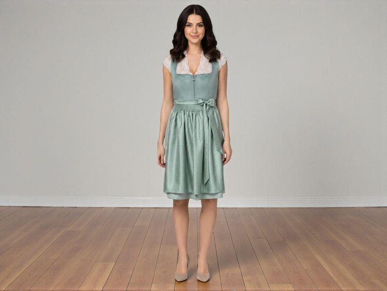 K&ouml;nnte beinhalten: Ein knielanges Dirndl in Salbeigr&uuml;n mit strukturiertem Stoff, Frontrei&szlig;verschluss und einer wei&szlig;en Spitzenbluse darunter. Das Kleid enth&auml;lt eine passende salbeigr&uuml;ne Sch&uuml;rze, die mit einer Schleife in der Taille gebunden ist.