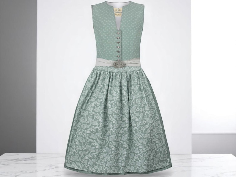 May include: A sleeveless, sage green dirndl dress with a floral pattern. The bodice has a V-neck, decorative buttons, and a decorative brooch. A white sash is cinched at the waist. The dress is a traditional style.