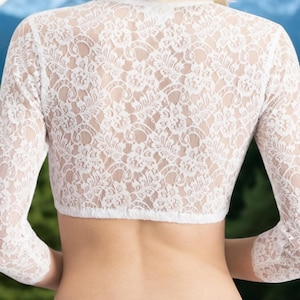 May include: White lace bolero with three-quarter sleeves. The delicate floral pattern of the lace is visible. The bolero is a cropped style, designed to be worn over other garments. The background is blurred, with green foliage and a mountain range.