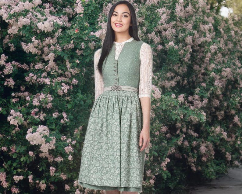 May include: A woman wearing a traditional green dirndl dress with a white blouse and lace sleeves. The dress features a floral pattern on the skirt and a decorative belt. The background is a lush display of pink flowers.