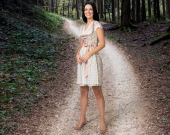 May include: A woman wearing a traditional white and beige dress with pink accents, standing on a forest path. The dress features lace detailing and a pink ribbon. She is wearing matching pink shoes.