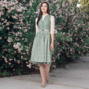 May include: A green dirndl dress with a floral pattern, featuring a button-down bodice and a full skirt. The dress includes a white lace blouse and a decorative belt. The woman is wearing beige heels and is standing in front of a flowering bush.