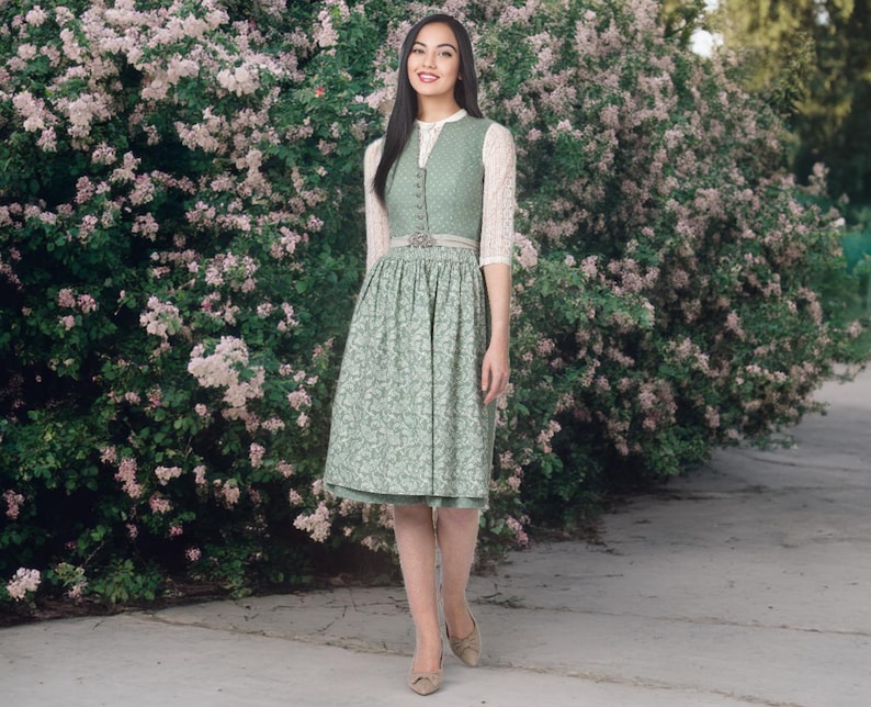 May include: A green dirndl dress with a floral pattern, featuring a button-down bodice and a full skirt. The dress includes a white lace blouse and a decorative belt. The woman is wearing beige heels and is standing in front of a flowering bush.