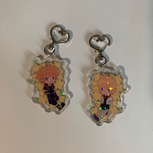Puede incluir: Dos llaveros de acrílico transparente con un anillo de metal en forma de corazón. Cada llavero presenta un personaje de dibujos animados con cabello rubio y un diseño de estrellas amarillas.