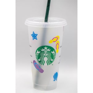 May include: A clear plastic reusable cup with a green Starbucks logo and various colourful beach-themed stickers, including flip flops, a seashell, and a star. The cup has a black straw.