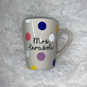May include: White ceramic mug with a polka dot pattern in pink, blue, yellow, and purple. The mug has a black inscription that reads "Mrs. LeRasoli" with silver glitter accents.