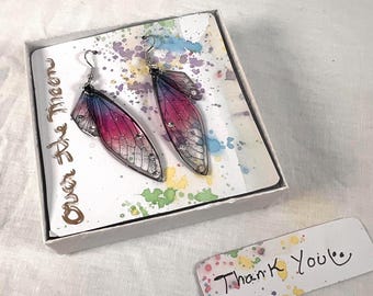 May include: A pair of iridescent pink and blue fairy wing earrings with silver accents. The earrings are packaged in a white box with a colorful background and the words "Over the Moon". A separate card with the words "Thank You" is also included.