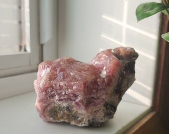 May include: A large, pink and white crystal with a rough, textured surface. The crystal is sitting on a white surface with a wooden edge.