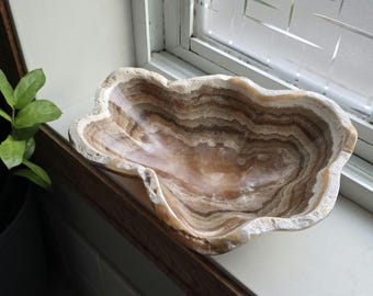 May include: A decorative, cloud-shaped bowl made of layered, natural stone. The bowl features bands of tan, brown, and cream colors, with a polished interior. The rim has a textured, rough appearance.
