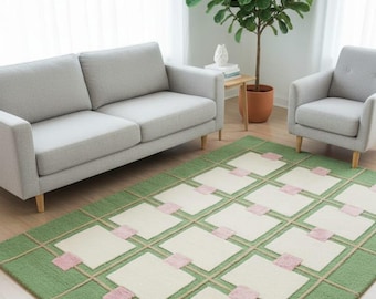 May include: A green and cream-colored area rug with a geometric pattern. The rug features a grid design with cream-colored squares, pink accents, and a green border. The rug is placed in a living room setting with a sofa and armchair.