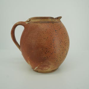 Puede incluir: Una jarra de cerámica redonda y bulbosa con un esmalte moteado en tonos terracota y bronceado. El recipiente cuenta con un asa curva en el lado izquierdo, un cuello corto y un pequeño pico vertedor en el borde, con una textura rústica.