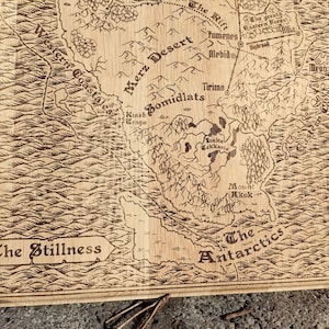 May include: A laser-engraved wooden map features a fantasy world with names like Merz Desert, Bomidlats, and The Antarctica.  The map's style is reminiscent of antique cartography, with detailed coastlines and landmasses.  The color palette is monochromatic, showcasing various shades of brown on the wood.  The text is clearly legible, adding to the map's intricate detail.  The map's overall aesthetic is rustic and charming, suitable for tabletop gaming or home decor.