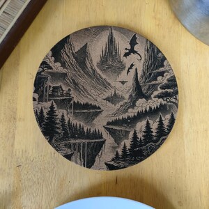 Può includere: Un sottobicchiere rotondo in sughero con un'illustrazione in bianco e nero di un paesaggio fantastico. L'illustrazione presenta una città galleggiante, montagne, alberi e un drago.
