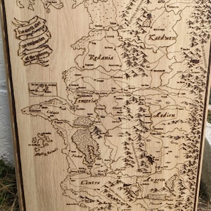 Può includere: Una mappa del mondo immaginario di The Witcher bruciata sul legno, con i regni di Redania, Kaedwen, Aedirn, Cintra e Angren. La mappa è etichettata con i nomi di vari luoghi e caratteristiche, tra cui montagne, fiumi e foreste.