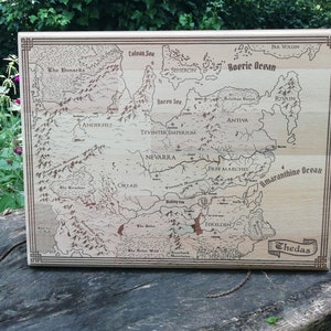 May include: A light brown wooden Thedas map features engraved lines and text, depicting various regions including Ferelden, Nevarra, and the Tevinter Imperium.  The map's border is a darker brown, and the names of the regions and bodies of water are clearly visible. The map is a decorative piece, ideal for fantasy fans and tabletop gamers.