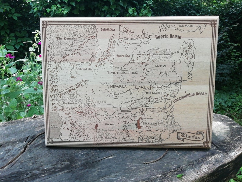 May include: A light brown wooden Thedas map features engraved lines and text, depicting various regions including Ferelden, Nevarra, and the Tevinter Imperium.  The map's border is a darker brown, and the names of the regions and bodies of water are clearly visible. The map is a decorative piece, ideal for fantasy fans and tabletop gamers.