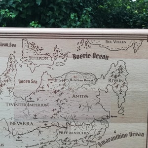 May include: A light brown wooden engraved map of a fantasy world. The map features various geographical features such as oceans, seas, mountains, forests, and countries with names like 'Seheron,' 'Antiiva,' 'Par Vollen,' 'Tevinter Imperium,' and 'Free Marches.' The map has a decorative border and is a unique piece for tabletop gaming or home decor.