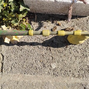 May include: A wooden wand with green and brown paint and yellow accents. The wand is lying on a grey concrete surface.