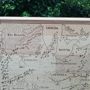 May include: A light brown wooden map of Thedas, from the Dragon Age video game series.  The map features engraved details including mountains, rivers, seas, and various regions such as The Donarks, Anderfels, Tevinter Imperium, Nevarra, and Free Marches.  The names of the regions are clearly labeled. The map has a decorative border. This laser-engraved wooden map is a collectible item for fans of fantasy and role-playing games.
