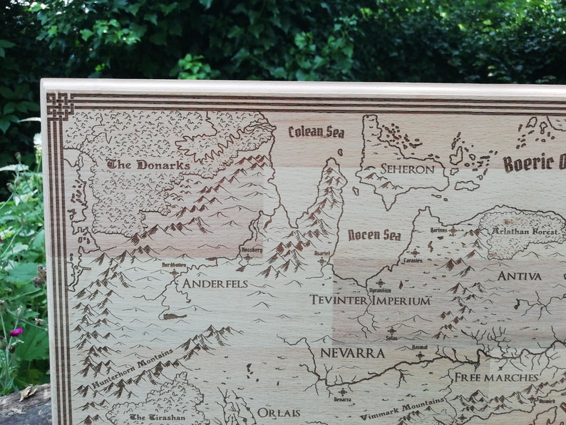 May include: A light brown wooden map of Thedas, from the Dragon Age video game series.  The map features engraved details including mountains, rivers, seas, and various regions such as The Donarks, Anderfels, Tevinter Imperium, Nevarra, and Free Marches.  The names of the regions are clearly labeled. The map has a decorative border. This laser-engraved wooden map is a collectible item for fans of fantasy and role-playing games.