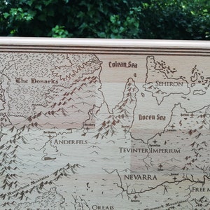 May include: A laser-engraved wooden map features various regions, including The Donarks, Anderfels, Seheron, Antiva, Tevinter Imperium, and Nevarra.  The map is bordered by a decorative frame and showcases detailed geographical features like mountains and seas, labeled with names like Colean Sea and Rocen Sea. The light-brown wood provides a natural contrast to the darker brown engraved lines.  The style evokes fantasy or role-playing game settings.