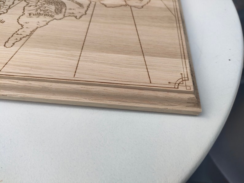 May include: Close-up view of a light brown wooden board with a laser-engraved map. The map features lines, text including 'Fulbrose', and decorative corner details. The board has a light wood grain and a simple frame. The image shows the bottom right corner of the board.