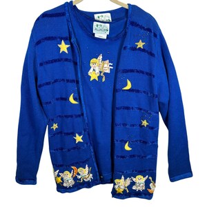 May include: A royal blue knit cardigan and matching tank top set featuring horizontal sequined stripes, yellow star and crescent moon appliques, and embroidered angel figures. The garments include white labels with green text and graphics.