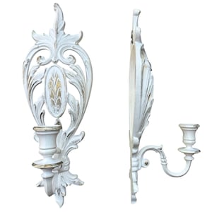 May include: A pair of wall-mounted candle sconces featuring an ornate, white-painted frame with distressed gold accents. The design includes intricate scrollwork and a central oval medallion, with a single candle cup extending from the base.