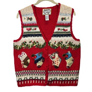 May include: A red knit vest featuring a V-neckline and a button-front closure. The garment displays a festive pattern with embroidered holly, poinsettias, and four decorative stockings. The label reads Heirloom Collectibles with care instructions.