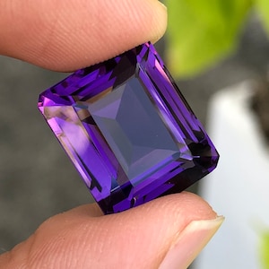 Peut inclure: Une pierre précieuse d'améthyste violette foncée taillée en forme rectangulaire avec des facettes. La pierre précieuse est tenue dans une main.