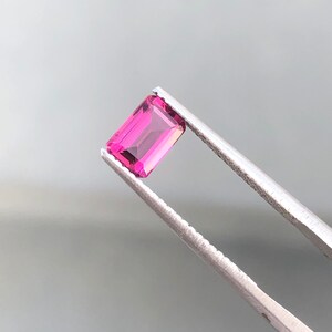 May include: A pink rectangular gemstone held by tweezers. The gemstone is cut into a rectangular shape with facets.