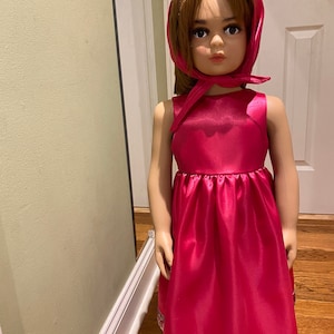 May include: A pink satin dress with a floral print trim. The dress has a sleeveless bodice and a full skirt. The model is wearing a matching pink satin head covering.