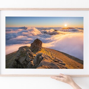 May include: A panoramic view of a mountain peak with a rocky outcrop, surrounded by a sea of clouds at sunset. The sun is setting behind the clouds, casting a warm glow over the scene.