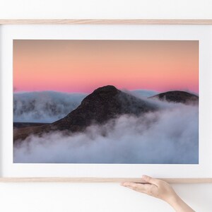 Könnte beinhalten: Ein Berggipfel ragt bei Sonnenuntergang über einem Meer aus Wolken empor. Der Himmel ist in leuchtenden Rosa- und Orangetönen gefärbt, mit zarten Wolken, die am Horizont schweben.