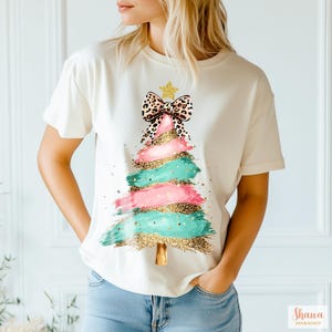 Op de afbeelding: Een cr&egrave;mekleurig T-shirt met korte mouwen en een afbeelding van een kerstboom gemaakt van horizontale penseelstreken in roze, groenblauw en goudglitter. De boom is bekroond met een gouden ster en een grote strik met luipaardprint.