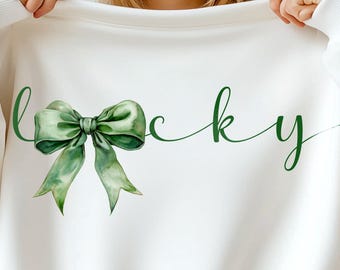 May include: A white long-sleeve sweatshirt featuring a green watercolor-style bow graphic that replaces the letter L in the word lucky, written in a dark green cursive script font across the chest of the garment.