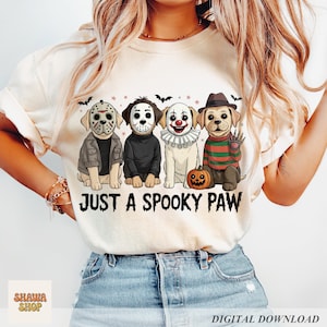 May include: A cream-colored t-shirt featuring a graphic of four dogs dressed as horror movie characters, including masks and costumes. Below the dogs, the text reads JUST A SPOOKY PAW in a black, distressed font. The design includes small bats and stars.