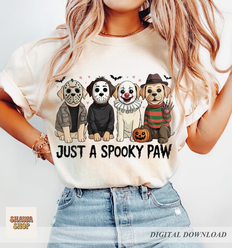 May include: A cream-colored t-shirt featuring a graphic of four dogs dressed as horror movie characters, including masks and costumes. Below the dogs, the text reads JUST A SPOOKY PAW in a black, distressed font. The design includes small bats and stars.