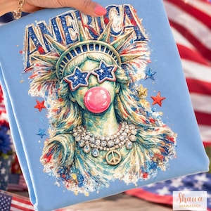 May include: A light blue sweatshirt featuring a graphic of the Statue of Liberty wearing star-shaped sunglasses and blowing a pink bubble. The word AMERICA is printed above in blue, with red and blue stars and a peace sign necklace accenting the design.