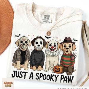 May include: A cream-colored t-shirt featuring a graphic of four dogs dressed as horror movie characters. The text below reads JUST A SPOOKY PAW. The dogs are wearing masks and costumes, including a hockey mask, clown makeup, and a striped sweater.