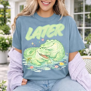 May include: A dusty blue t-shirt featuring a graphic of two light green alligators in water. The top alligator rests its chin on the bottom one. The bottom alligator wears a yellow bow. The text LATER is printed in large, light green letters above.