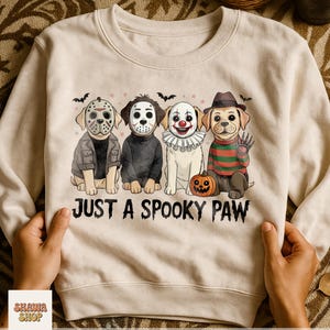 May include: A beige crewneck sweatshirt featuring a graphic of four dogs dressed as horror movie characters. The text "JUST A SPOOKY PAW" is printed below the dogs. The illustration includes bats, stars, and a small pumpkin on the ground.