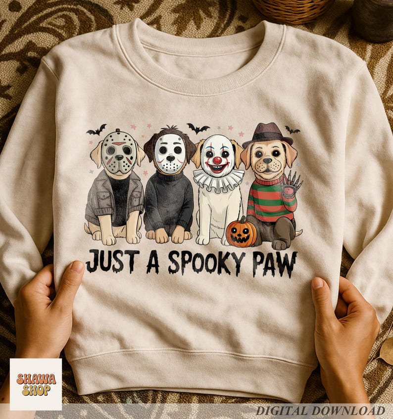 May include: A beige crewneck sweatshirt featuring a graphic of four dogs dressed as horror movie characters. The text "JUST A SPOOKY PAW" is printed below the dogs. The illustration includes bats, stars, and a small pumpkin on the ground.