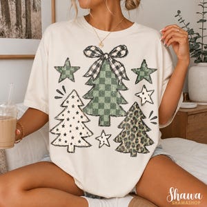May include: An off-white short-sleeve t-shirt featuring a graphic print of three stylized Christmas trees and stars. The central tree has a black and white buffalo plaid bow, a green checkered pattern, and a leopard print tree to the right.