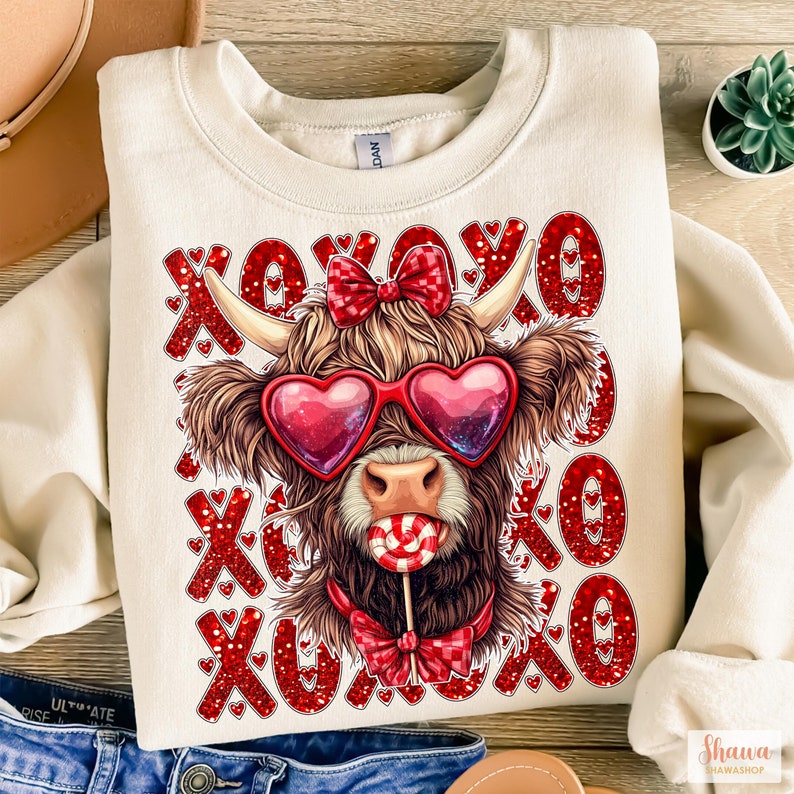 May include: A cream-colored sweatshirt with a red glitter XOXO pattern and a cartoon Highland cow wearing heart-shaped sunglasses and a red bow tie. The cow is holding a red and white lollipop.