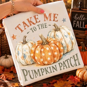 Può includere: Un articolo in tessuto bianco con un'illustrazione ad acquerello di tre zucche. La zucca centrale è arancione a pois bianchi, affiancata da due zucche dai toni blu. Il testo recita "TAKE ME TO THE PUMPKIN PATCH" in arancione e verde.